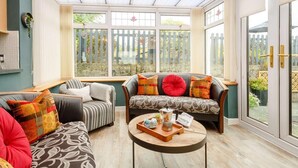A conservatory with a sofa set, a coffee table with a tray, and a view of a garden through large windows. - 2 Bed in Moelfre (oc-75013) (Moelfre)