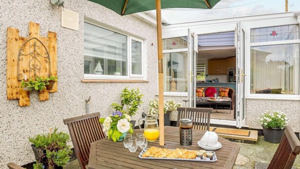 A patio with a wooden table, chairs, a green umbrella, and a breakfast setup including croissants and juice. - 2 Bed in Moelfre (oc-75013) (Moelfre)