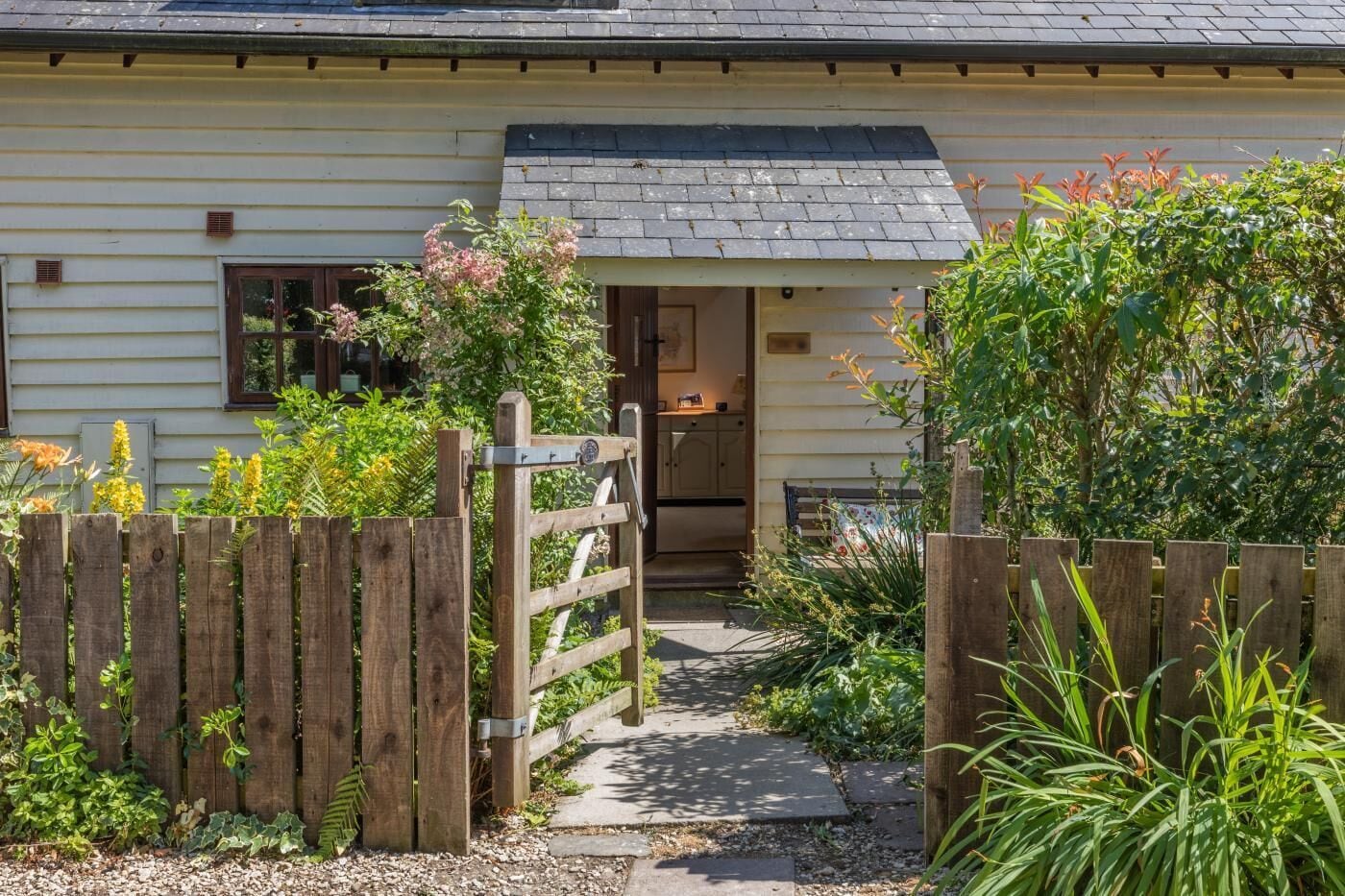 Une maison avec un portail en bois, un chemin menant à l’entrée et une variété de plantes qui ornent la propriété.