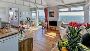 A spacious living area with a wooden floor, a dining table, a sofa, and a TV. There is a view of the sea through large windows. - 1 Bed in Torpoint (oc-74824) (Millbrook)