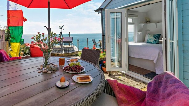 A deck with a table set for breakfast, a red umbrella, and a view of the sea. - 1 Bed in Torpoint (oc-74824) (Millbrook)