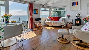 A spacious living area with a wooden floor, a white sofa, a rocking chair, and a dining table set for two. - 1 Bed in Torpoint (oc-74824) (Millbrook)