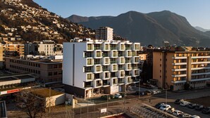 Un immeuble moderne à plusieurs étages, doté de balcons, entouré d’un parking et adossé à un paysage montagneux.