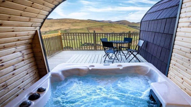 Ein Whirlpool mit einer Holzterrasse und Stühlen, umgeben von einem Holzzaun und Hügeln im Hintergrund.