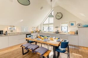 A modern kitchen with a dining area, featuring a wooden table, blue chairs, and a clock on the wall. - 4 Bed in Bideford (oc-75358) (Northam)