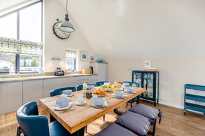 A modern kitchen with a wooden dining table set for breakfast, blue chairs, and a view of the street through large windows. - 4 Bed in Bideford (oc-75358) (Northam)