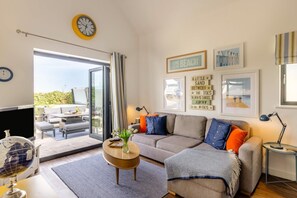 A living room with a sofa, a coffee table, and a television. There are framed pictures on the wall and a window with curtains. - 4 Bed in Bideford (oc-75358) (Northam)