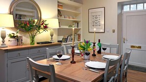 Een eetgedeelte met een houten tafel gedekt voor vier personen, omringd door stoelen. Daarboven hangt een spiegel, en tegen de muur is een boekenkast met boeken en decoratieve objecten te zien, evenals een lijst met een afbeelding.
