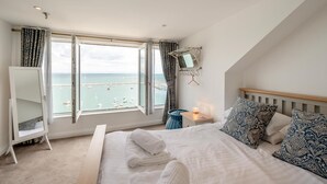 Ein Schlafzimmer mit Bett, Nachttisch mit Lampe, Kleiderschrank und einem großen Fenster mit Meerblick.