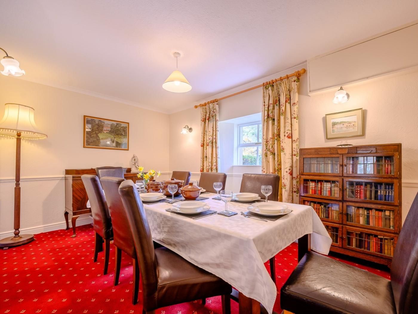 Une salle à manger avec une table dressée pour quatre personnes, entourée de fauteuils en cuir, d’une bibliothèque et d’une fenêtre agrémentée de rideaux.