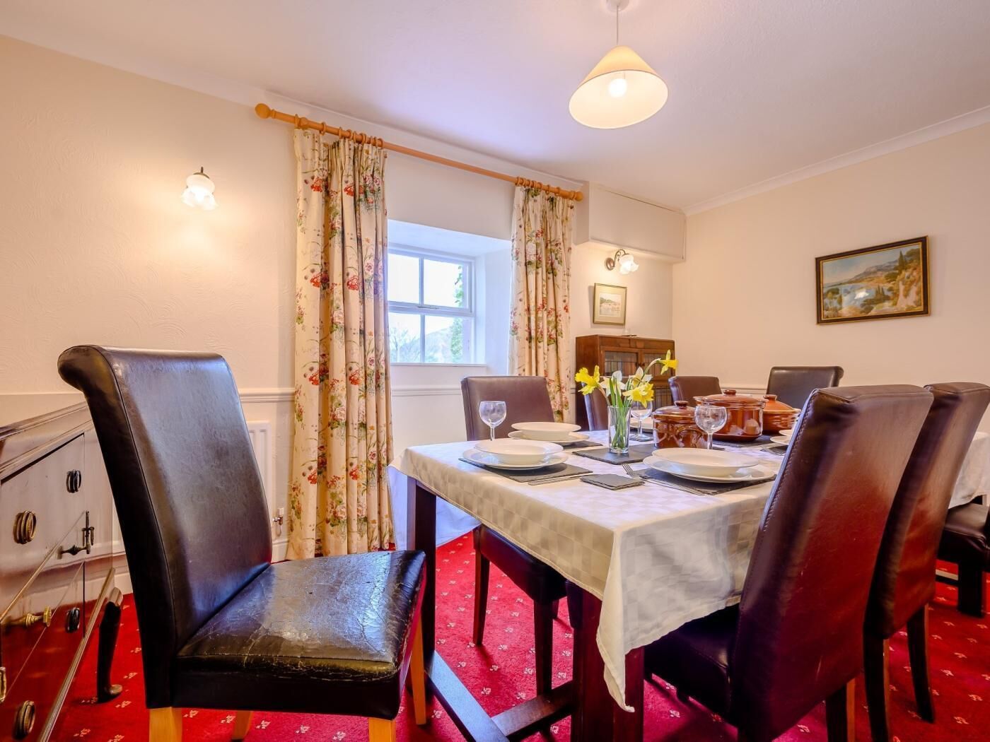 A dining room with a table set for six, six chairs, a vase with flowers, and a painting on the wall.