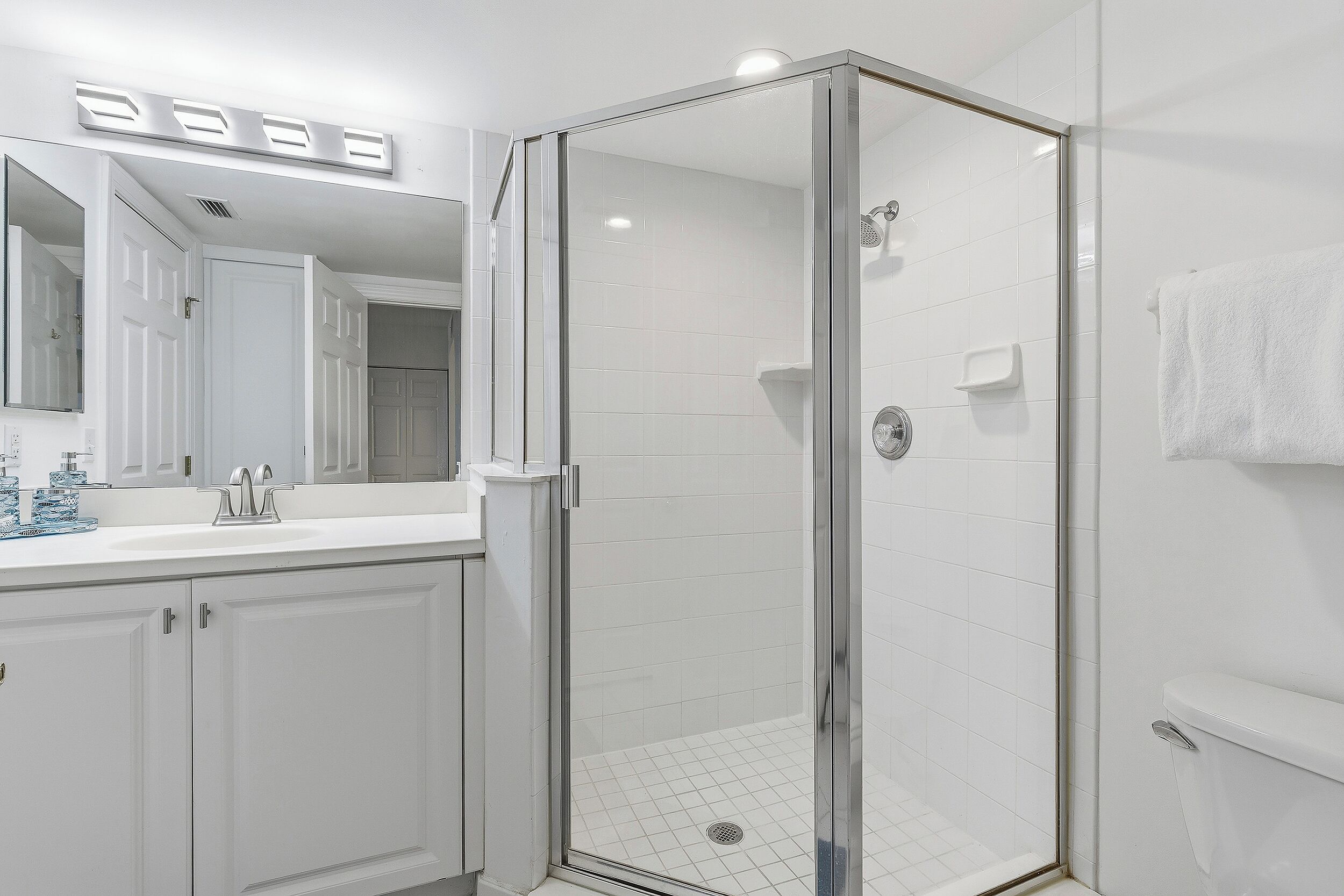 Un baño moderno con ducha envuelta en vidrio, azulejos blancos, un espejo grande y encimera blanca.