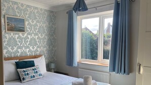 A bedroom with a bed, a window with curtains, a radiator, and a wall with a patterned wallpaper. - 2 Bed in Norwich (oc-75398) (Mundesley)
