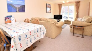 A living room with a sofa set, a dining table with a sailboat tablecloth, a television, and a window with curtains. - 2 Bed in Norwich (oc-75398) (Mundesley)