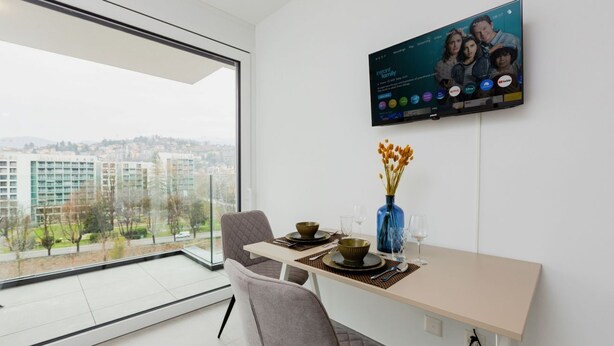 Un'area pranzo moderna con una grande finestra, una TV a schermo piatto montata a parete e un vaso con fiori sul tavolo.