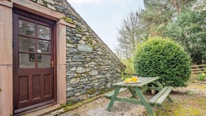 Ein Steinhaus mit einer Holztür, einer Steinsäule, einem grünen Picknicktisch mit einer Frucht- und Getränkeauswahl und einem Nadelbaum.