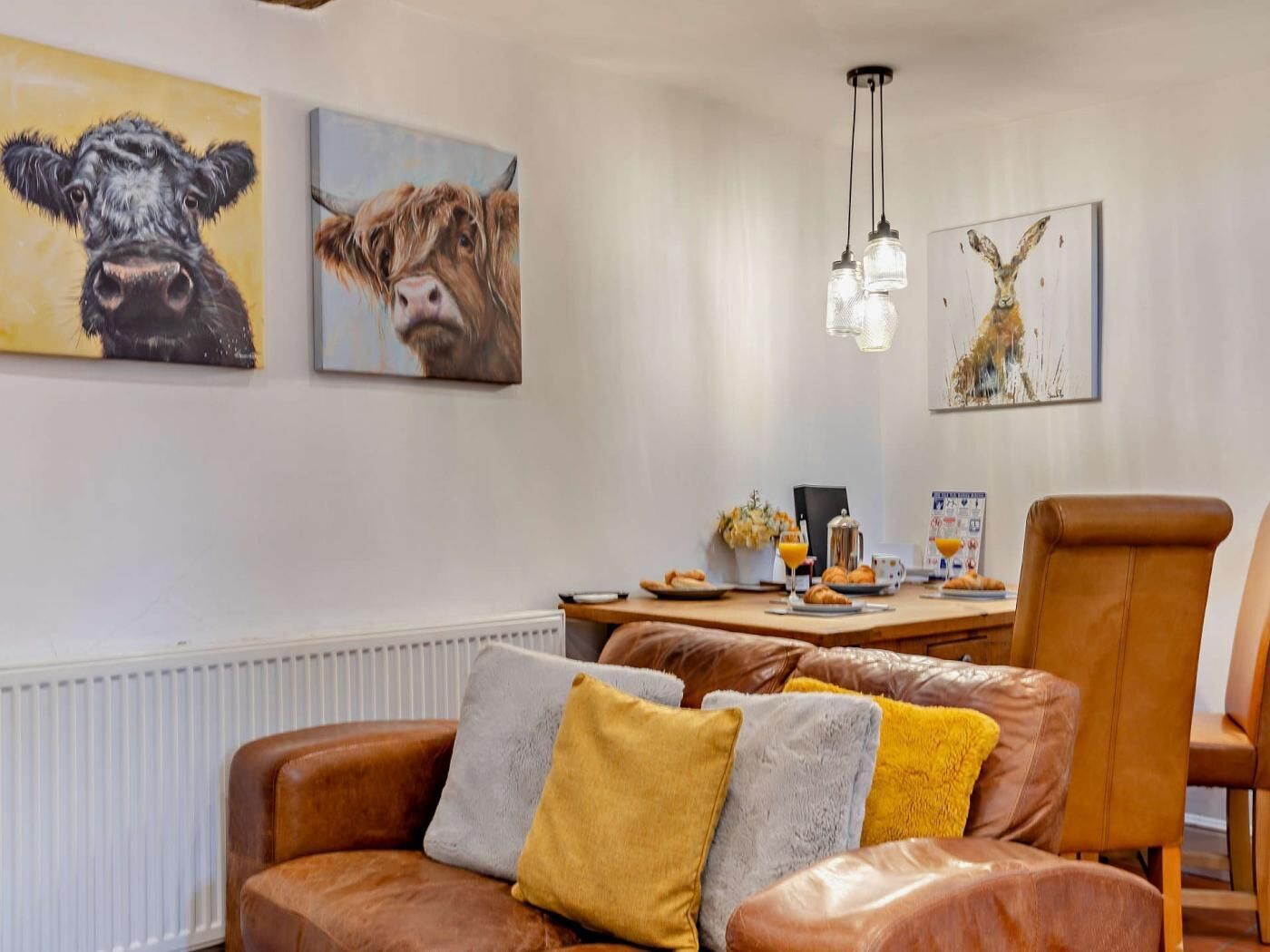 Een gezellige woonkamer met een bruine leren bank, een eettafel met ontbijtartikelen en kunstwerken aan de muren.