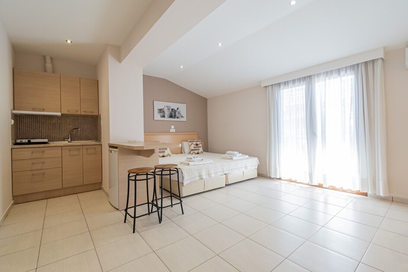 Een moderne hotelkamer met kitchenette, een bed met witte dekbedovertrek, een zithoekje en een groot raam met dunne gordijnen.