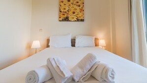 Un letto rifatto con lenzuola bianche e asciugamani arrotolati, due lampade sui comodini e un quadro di girasole al muro.