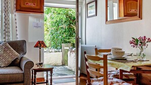 A cozy living room with a sofa, a small table with a lamp, a dining area with chairs and a table set for two, and a view of greenery outside. - 2 Bed in Keswick (oc-sz145) (Braithwaite)