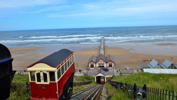 Ein Bahnhof am Meer mit einem roten Zug, Gleisen und einem Gebäude mit spitzer Dachform.