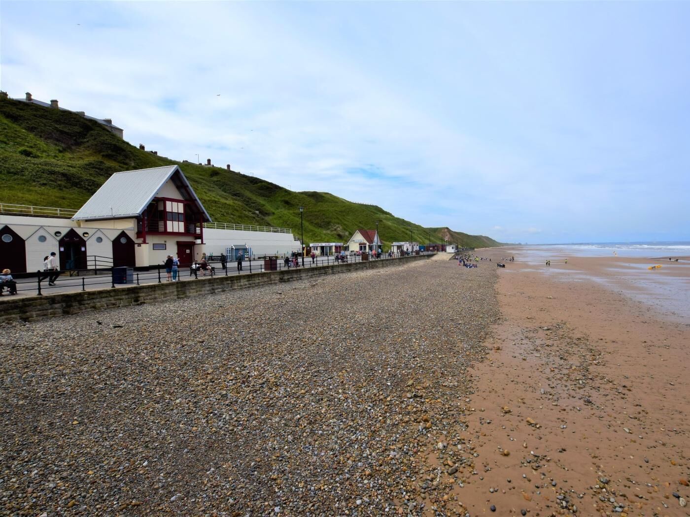Ein Kiesstrand mit einer Reihe von Gebäuden, darunter ein markantes rot-weißes Gebäude, und ein grasbewachsener Hügel im Hintergrund.