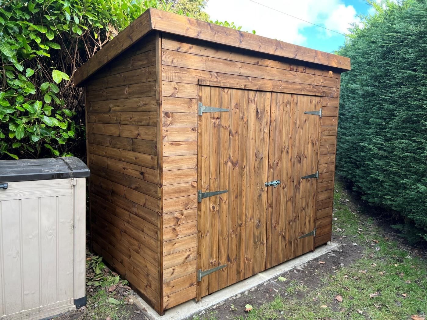 A wooden shed with a sloped roof, a door with a handle, and a small adjacent shed.