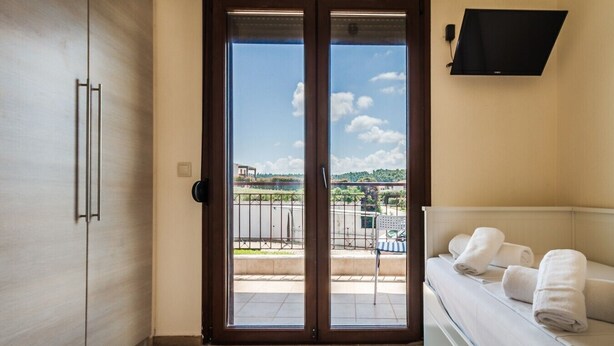 Un dormitori amb una porta corredissa de vidre a un balcó, un llit amb roba blanca, un televisor fixat a la paret i un armari de fusta.