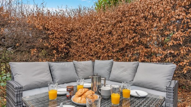 En fletsofa med grå puder, et bord med glasplade og morgenmadsprodukter, samt et baggrund af tørt løv.