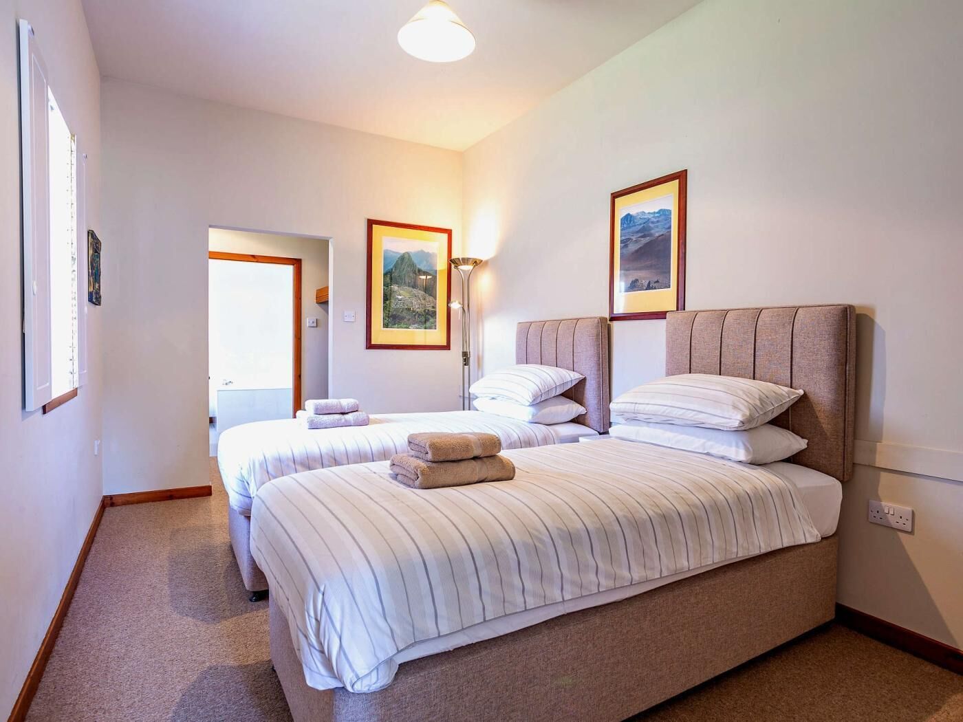 A hotel room with two beds, a standing lamp, and framed pictures on the walls.