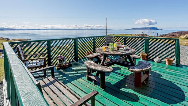 A deck with a view of a body of water and distant land, featuring outdoor seating and a table with items. - 3 Bed in Isle Of Skye (oc-ca071) (Aird of Sleat)