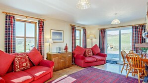 A room with a red sofa, a wooden cabinet, a dining table with chairs, and large windows with curtains. - 3 Bed in Isle Of Skye (oc-ca071) (Aird of Sleat)