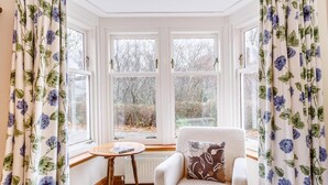 A room with a bay window, a chair, a small table, and curtains with a floral pattern. - 6 Bed in Lairg (oc-ca210) (Laxford Bridge)