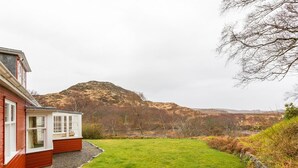 A red building with a white windowed porch, a green lawn, and a mountainous backdrop. - 6 Bed in Lairg (oc-ca210) (Laxford Bridge)
