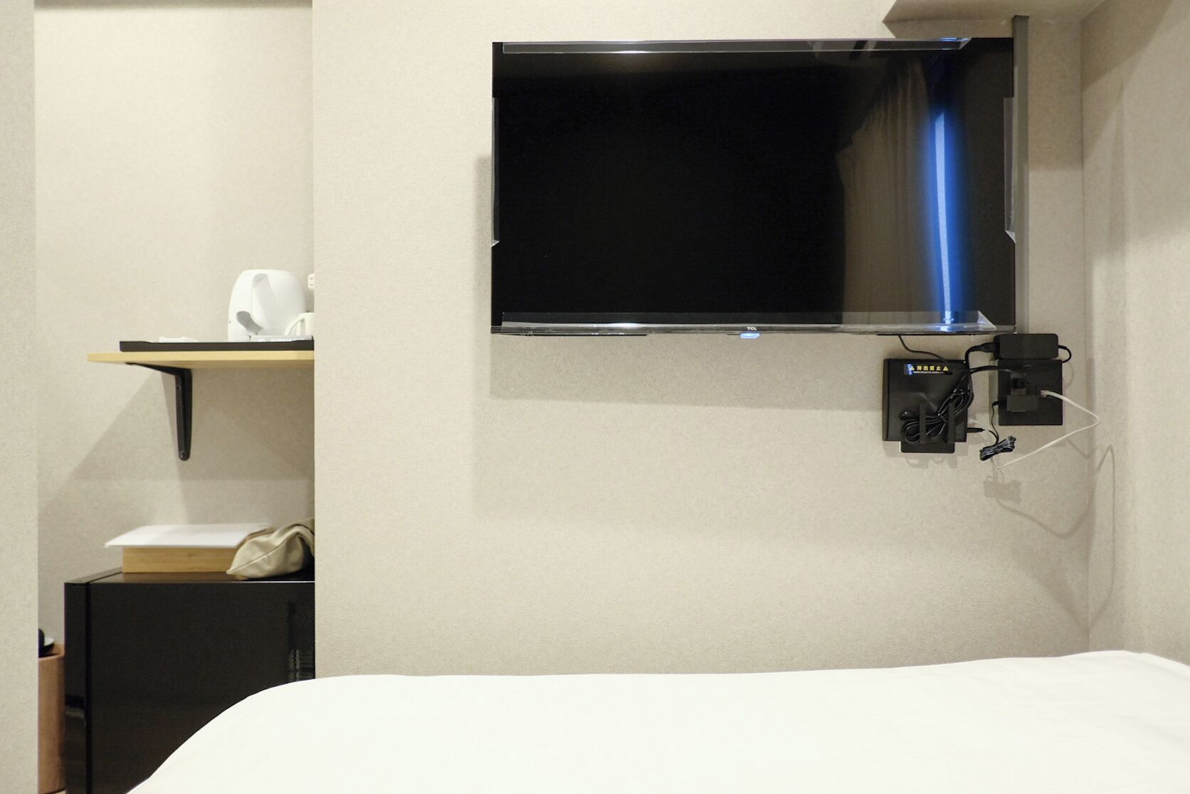Une chambre d’hôtel avec un lit, un téléviseur fixé au mur, une petite étagère avec une tasse et un livre, et une prise électrique avec une fiche.