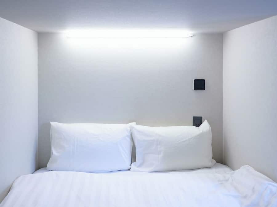 Uma cama de solteiro com roupa de cama branca num quarto com paredes e luz no teto brancas.