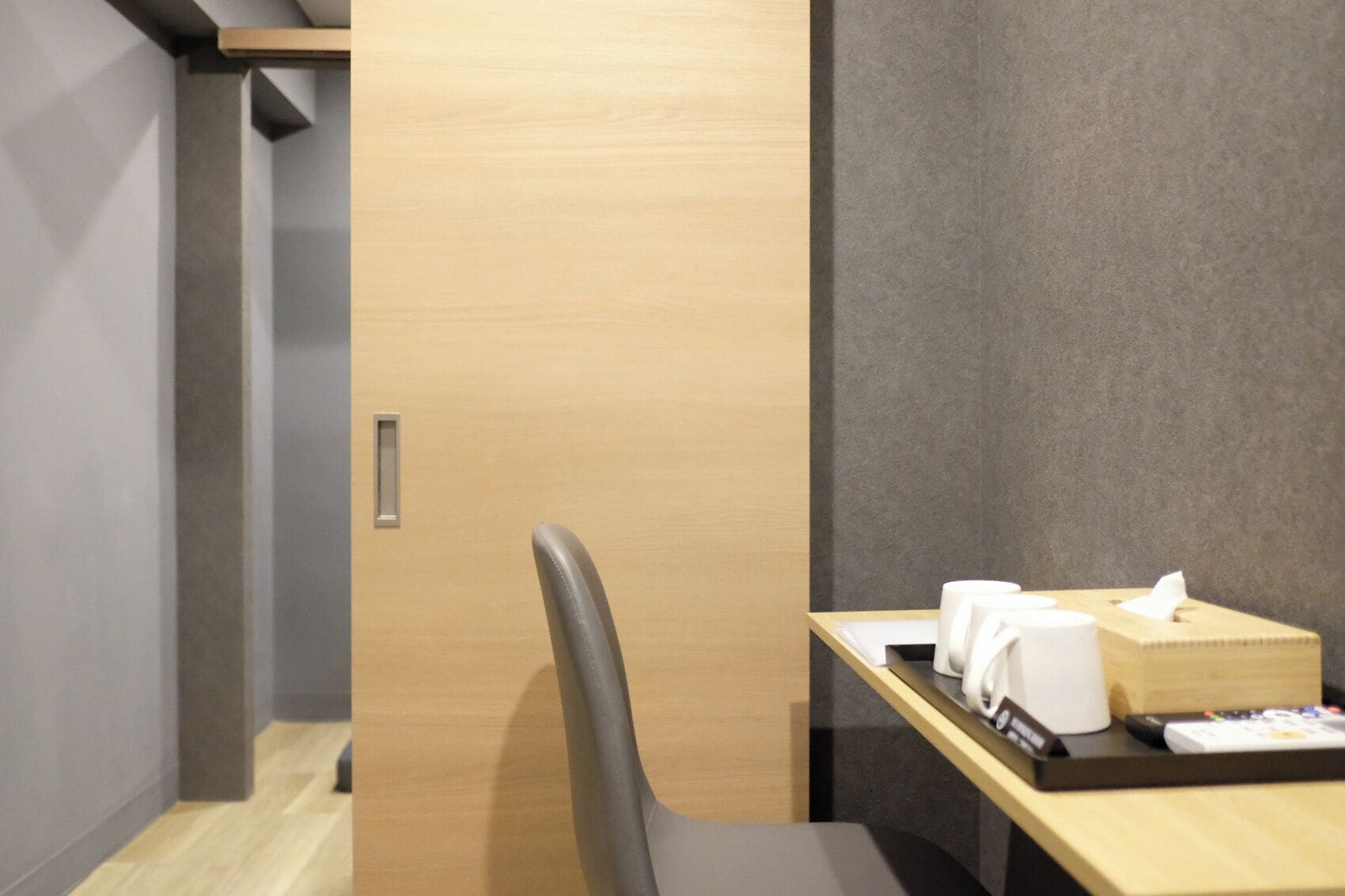 A hotel room with a wooden door, a desk with a tissue box, a remote control, and a chair.
