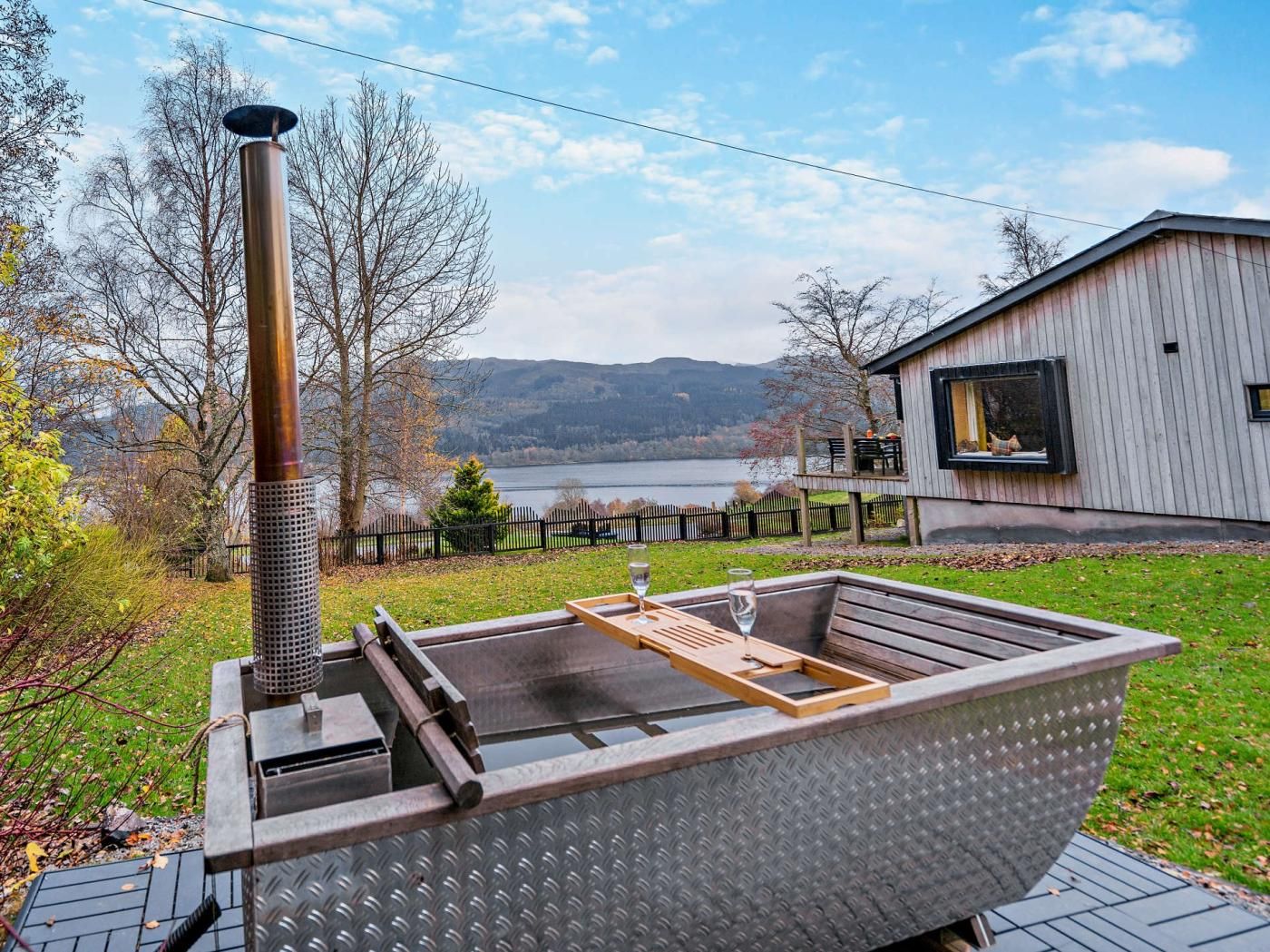 A sauna with a metal structure and a wooden bench, set against a backdrop of a lake and a wooden building.