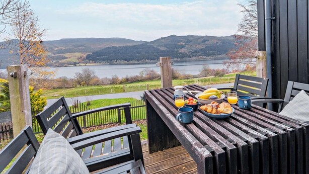A wooden deck with a table set for breakfast overlooking a lake and hills. - 3 Bed in Pitlochry (oc-ca207) (Tressait)