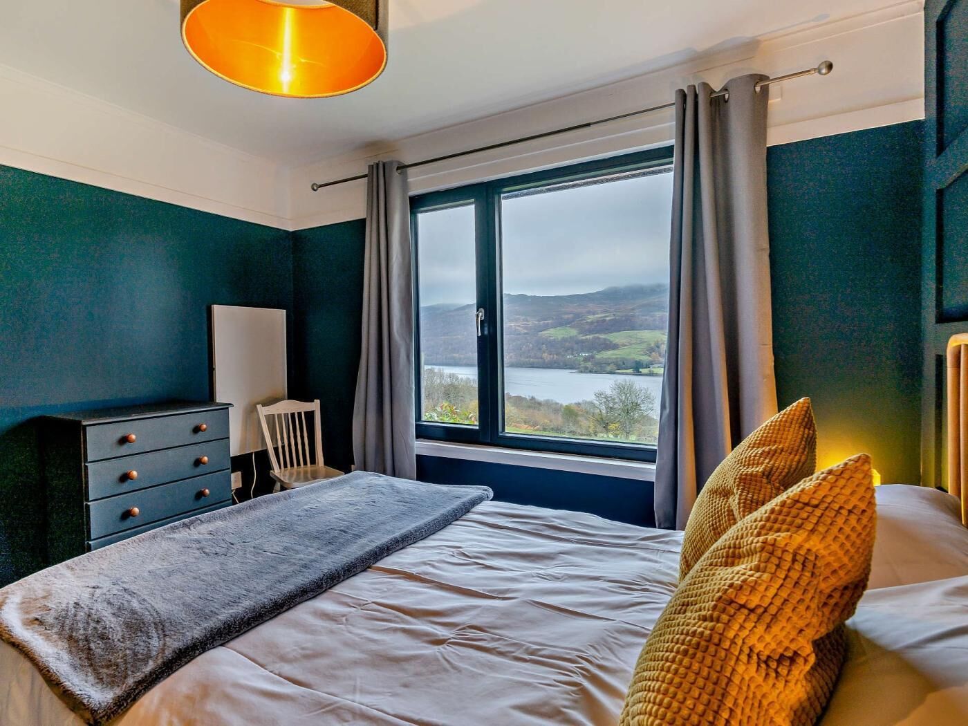 A bedroom with a bed, a dresser, a chair, a lamp, and a view of a lake and hills.