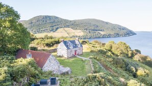 A stone house with a red door, surrounded by lush greenery and a view of a lake and hills. - 2 Bed in Drumnadrochit (oc-ca315) (Strone)