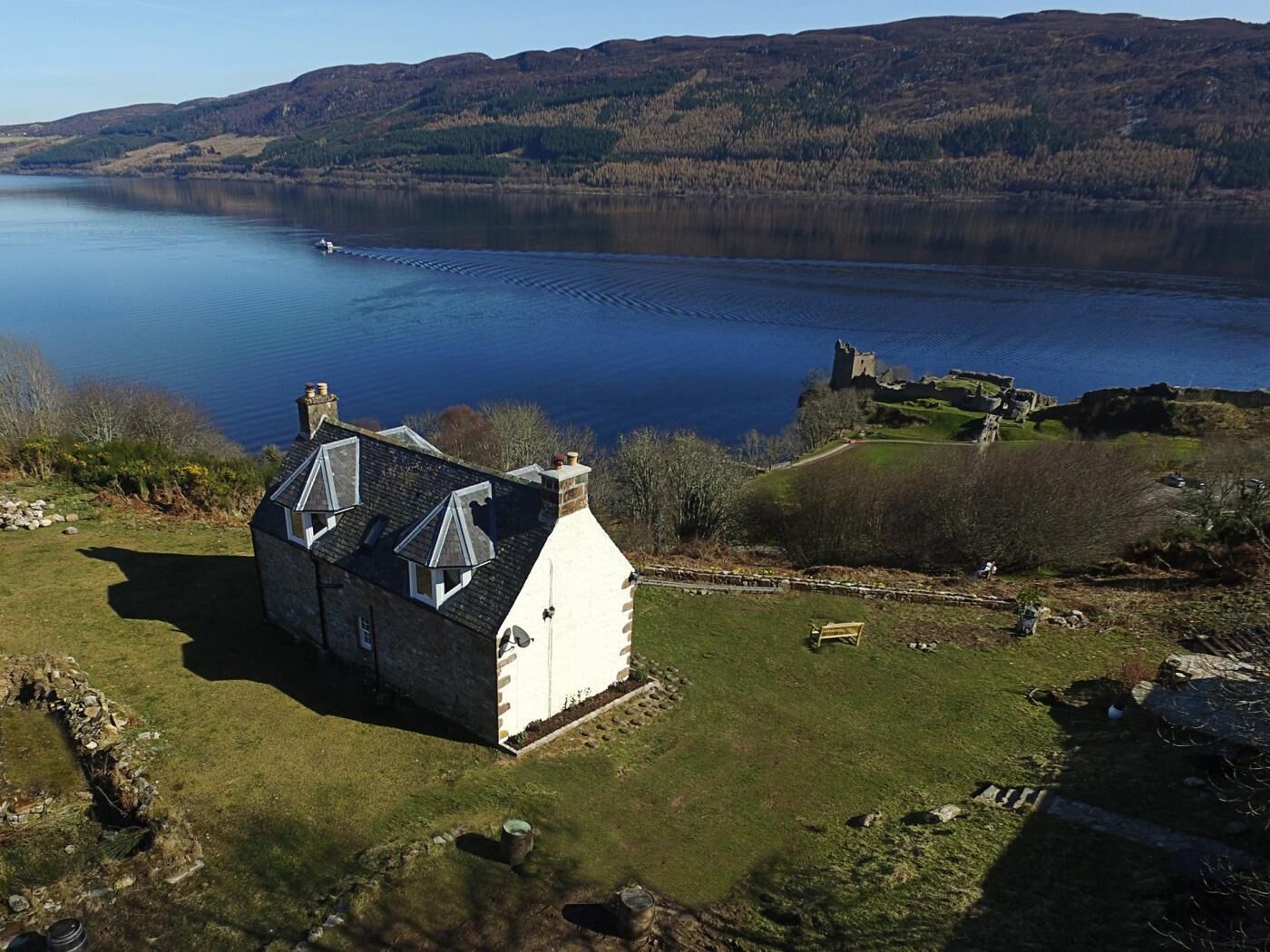 A stone house with a sloped roof and a chimney, situated on a grassy area with a view of a large body of water and a distant castle.