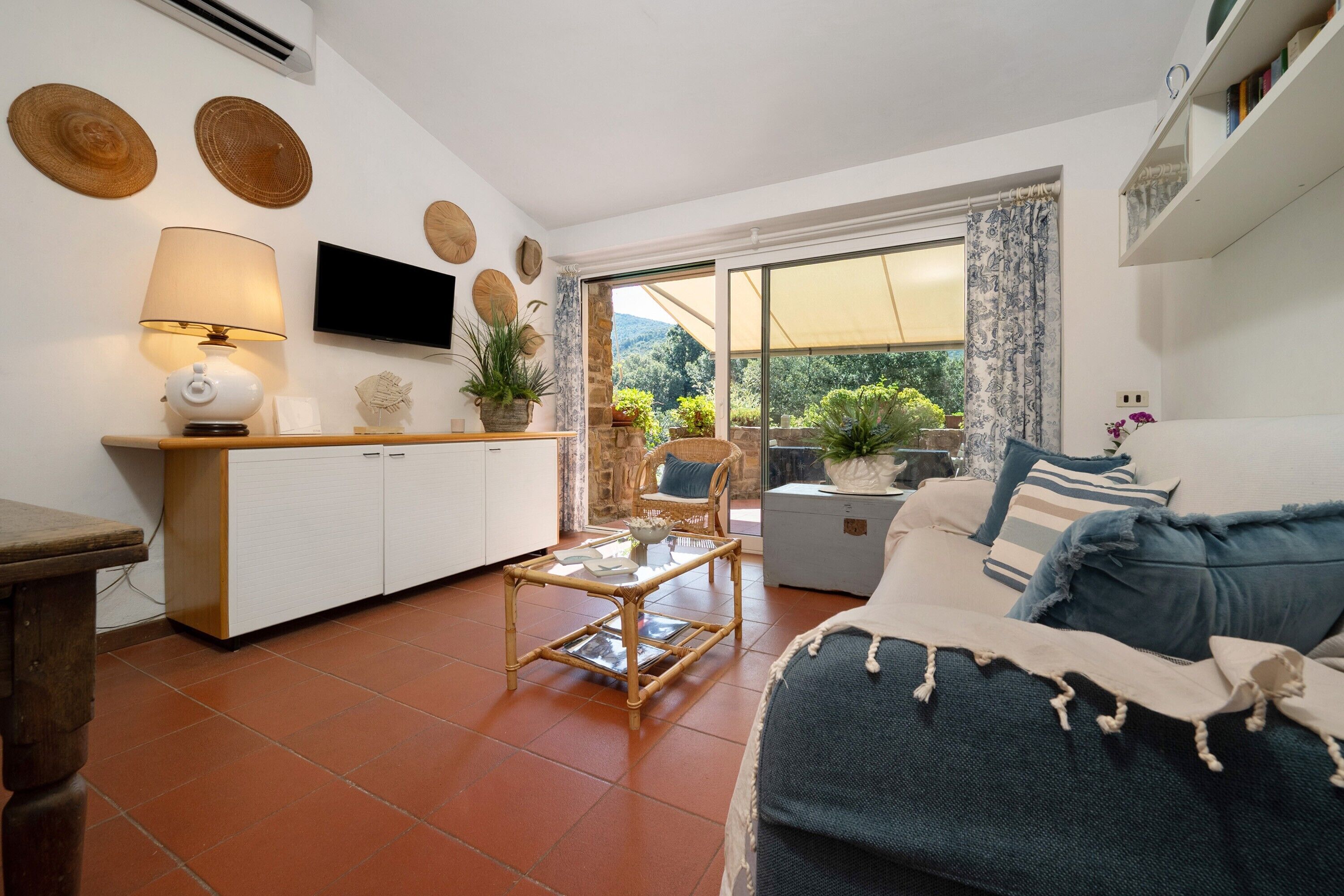 Una sala de estar acogedora con un sofá, una mesita de centro y un televisor. Hay una zona de cocina con un armario y una lámpara. La habitación tiene vistas al exterior a través de un ventanal grande.