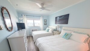 A hotel room with two beds, a ceiling fan, a large window with curtains, a flat-screen TV, and a painting on the wall. - Ocean view, steps to beach with a relaxing pool! (Ocean City)