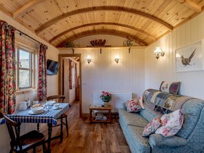 Ein gemütliches Zimmer mit Holzbalkendecke, einem Esstisch mit Stühlen, einer Couch mit floralen Kissen und einem an der Wand befestigten Fernseher.