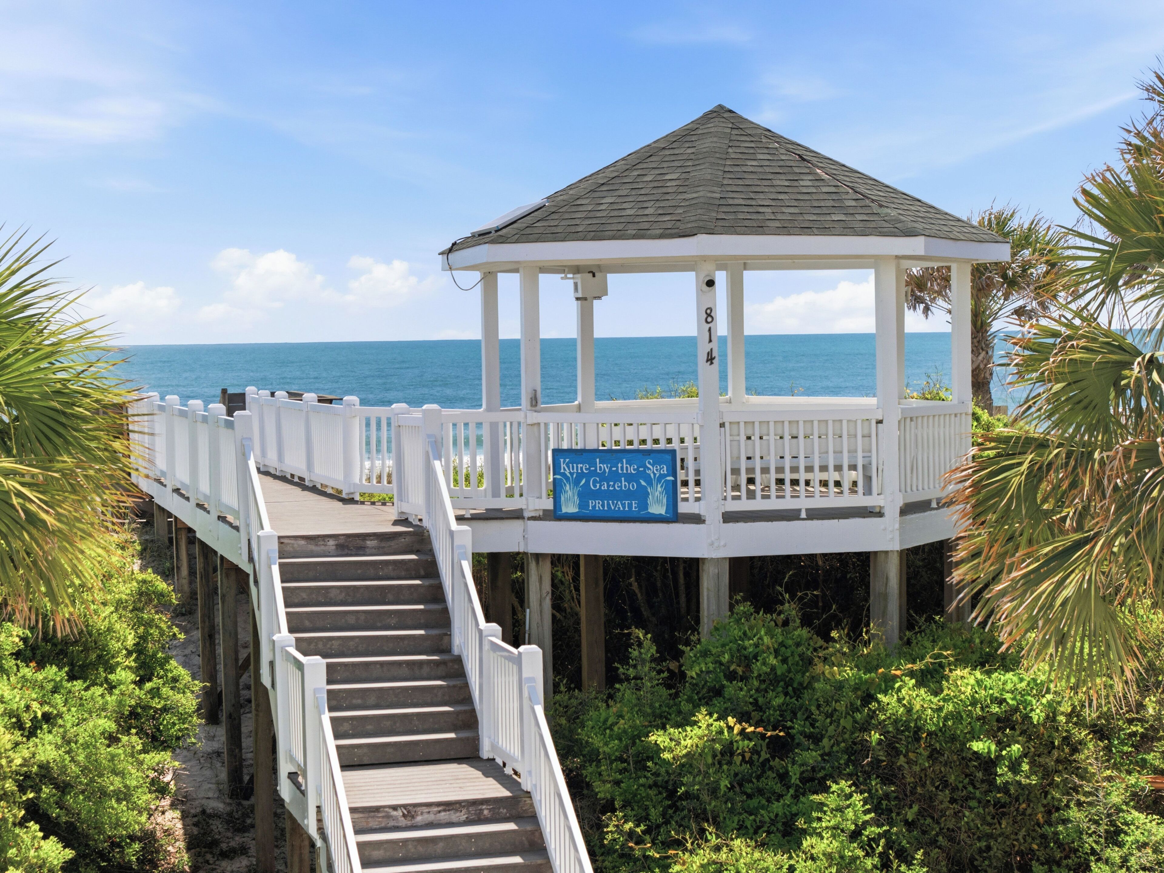 A gazebo with a sign reading 'Kure-by-the-Sea Gazebo PRIVATE' on a wooden pier with stairs leading to it, surrounded by greenery and overlooking the sea.