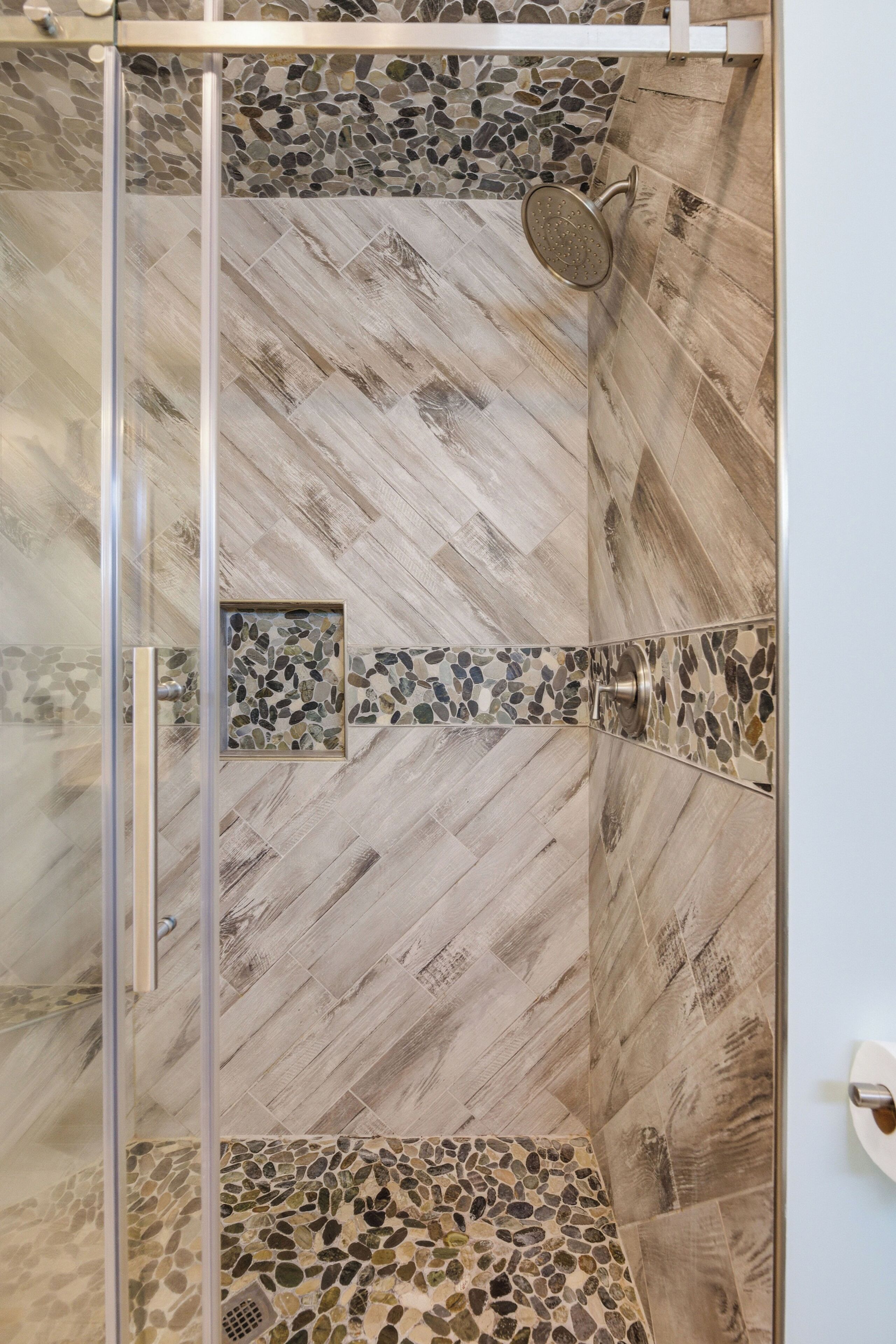Ducha con puerta de vidrio, pared de mosaico de piedra y suelo de madera en patrón de espiga.