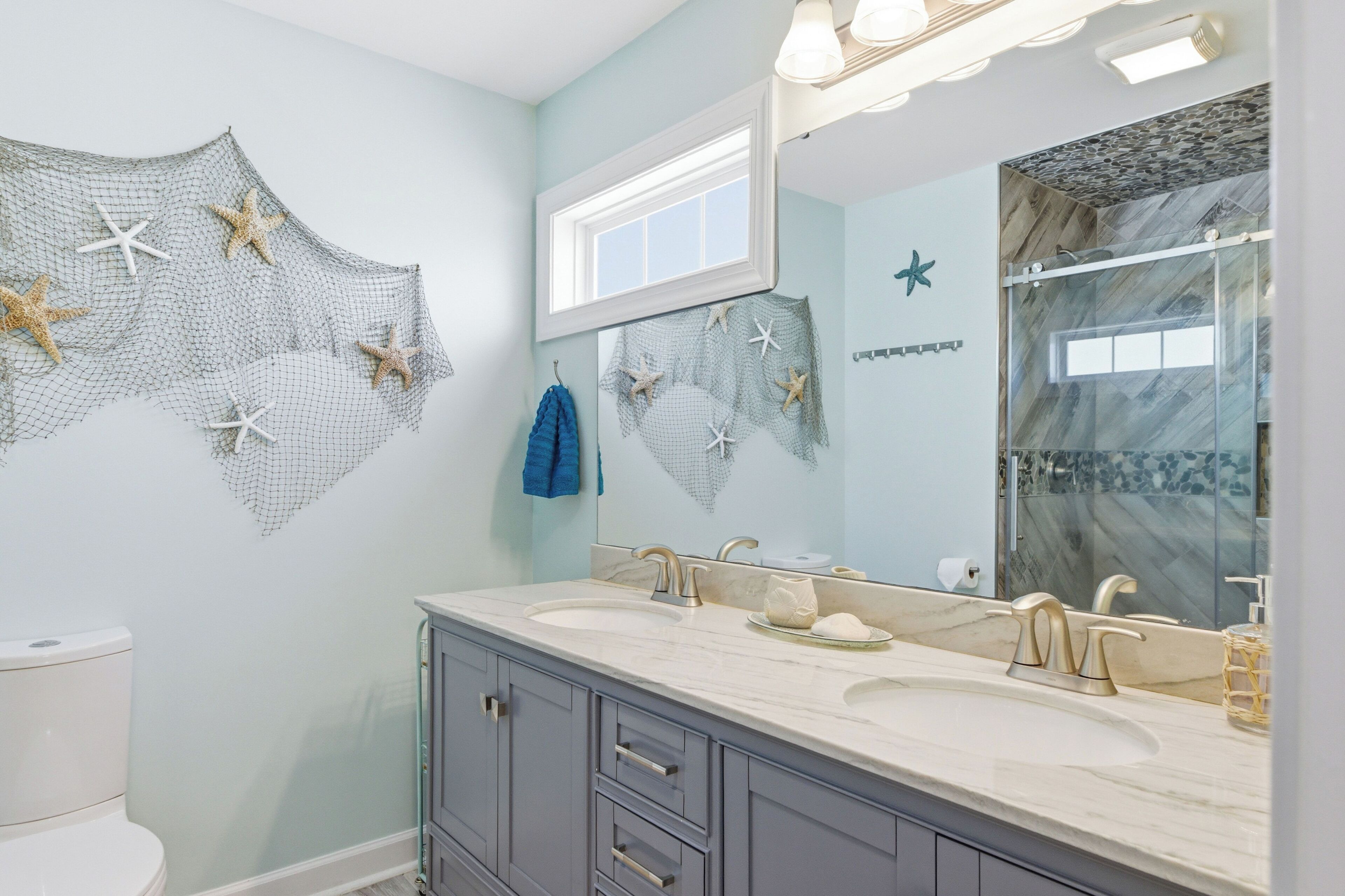 Un baño con un espejo grande, dos lavamanos, un inodoro y estrellas de mar decorativas en la pared.