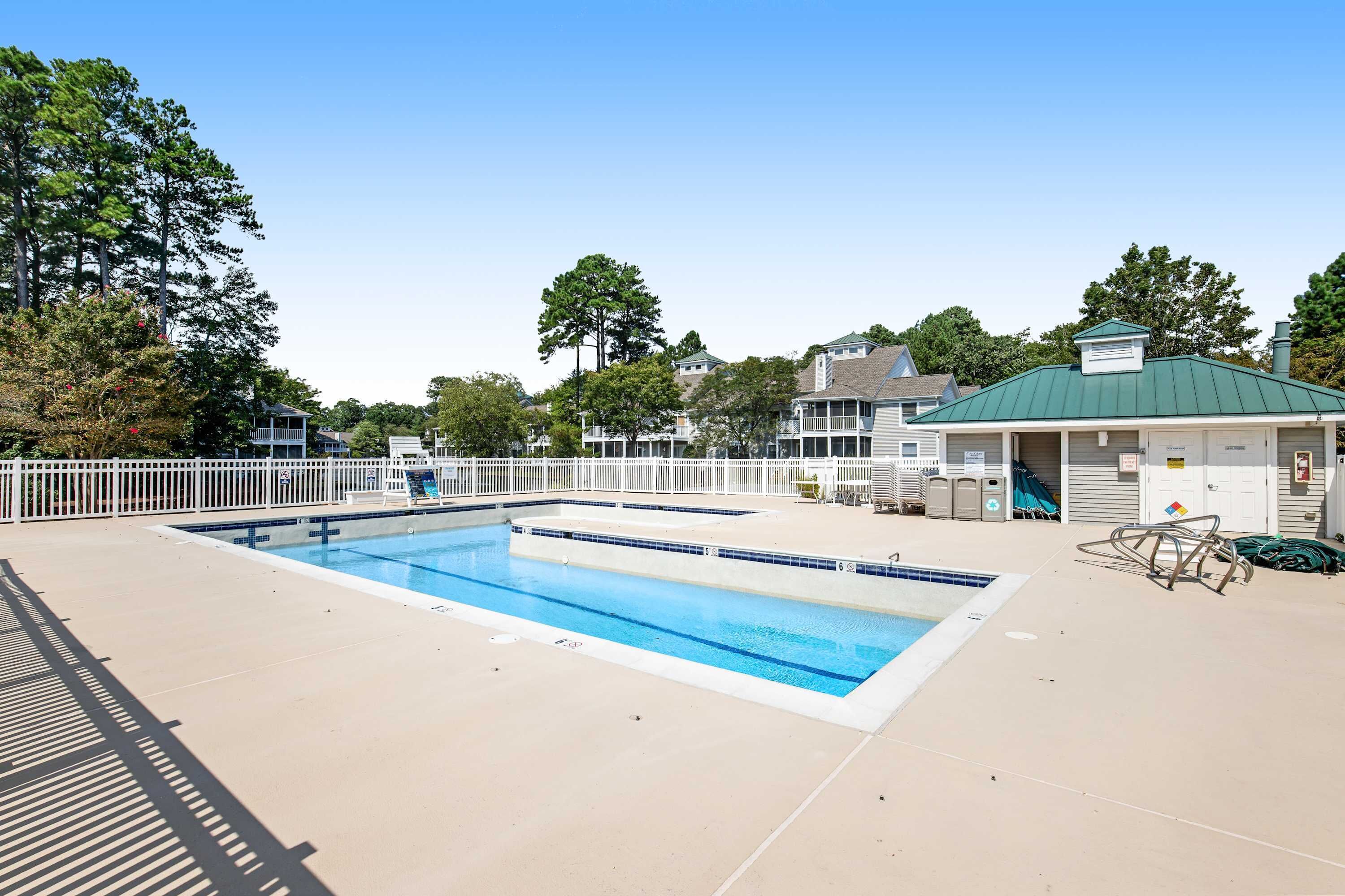 A swimming pool with a white fence, a covered area with a green roof, and trees in the background.