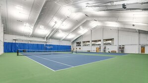 Una cancha de tenis interior con red, suelo azul y verde, y un techo alto con vigas y luminarias a la vista.